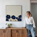 A woman in a white blouse and jeans stands by a wooden sideboard, admiring the Nightshade Canvas Art, which features large blue flowers on a cream background. A potted plant and books are neatly arranged on the sideboard.