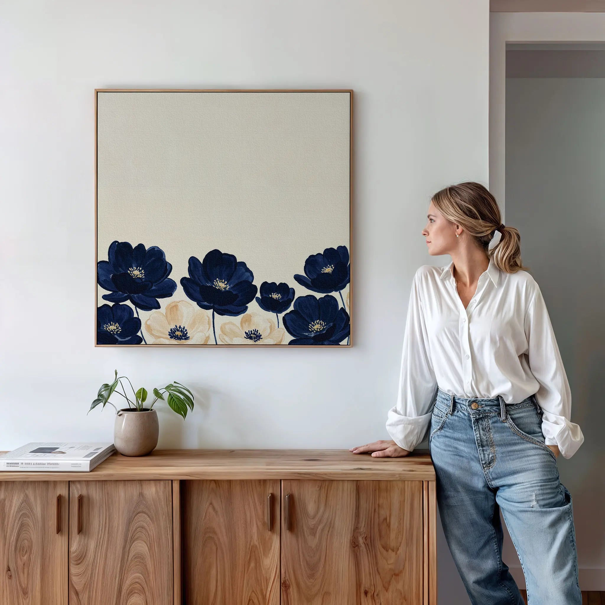 A woman in a white blouse and jeans stands by a wooden sideboard, admiring the Nightshade Canvas Art, which features large blue flowers on a cream background. A potted plant and books are neatly arranged on the sideboard.