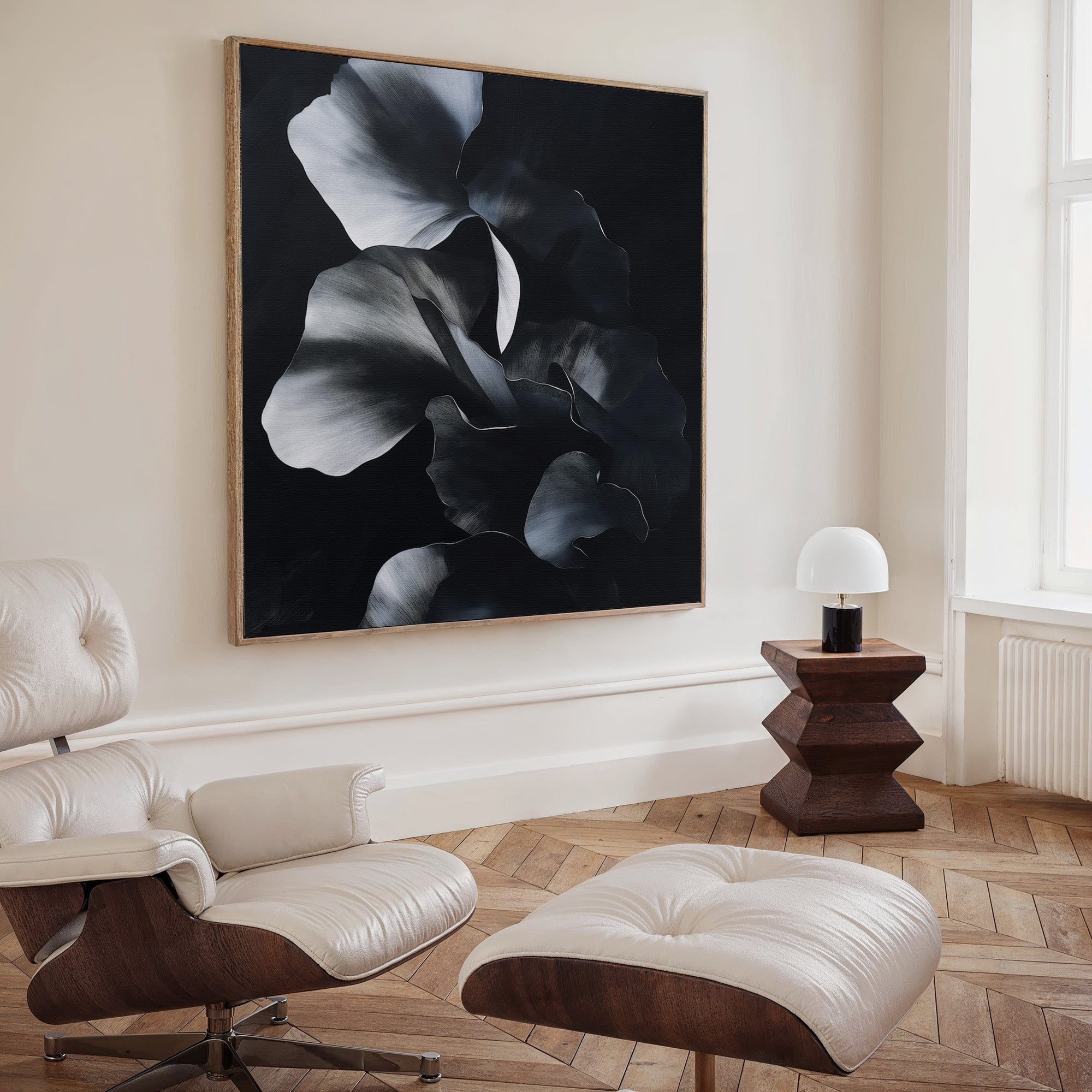 A modern living space highlights Nocturnal Bloom Abstract Canvas Art—a black-and-white floral painting—paired with a cream leather lounge chair, ottoman, geometric wooden side table, and white lamp by a tall window.