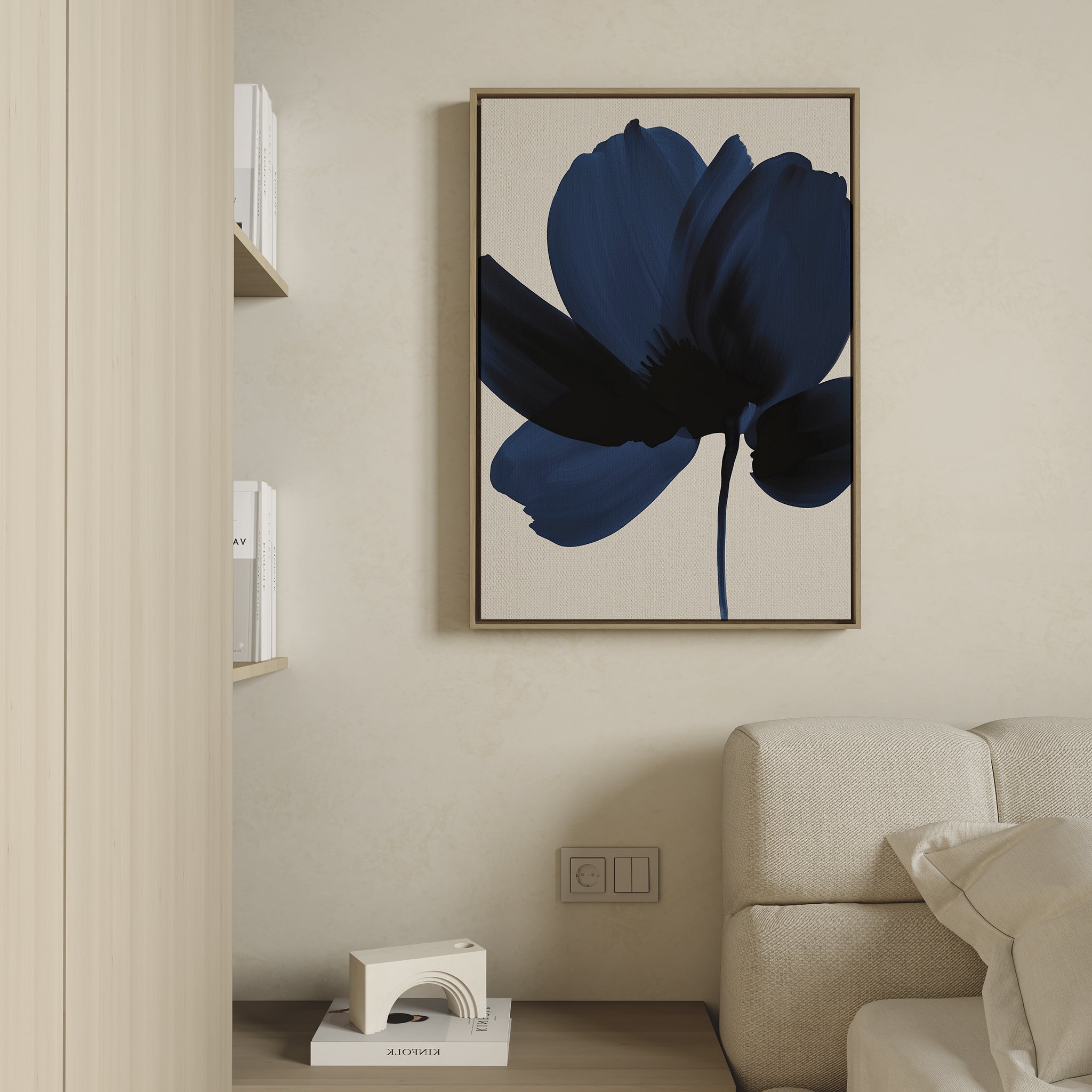 A minimalist living room showcases the Nocturne Blossom Canvas Art—a large framed abstract blue flower painting—on a beige wall above a light sofa, paired with a shelf holding books and decor for chic elegance.