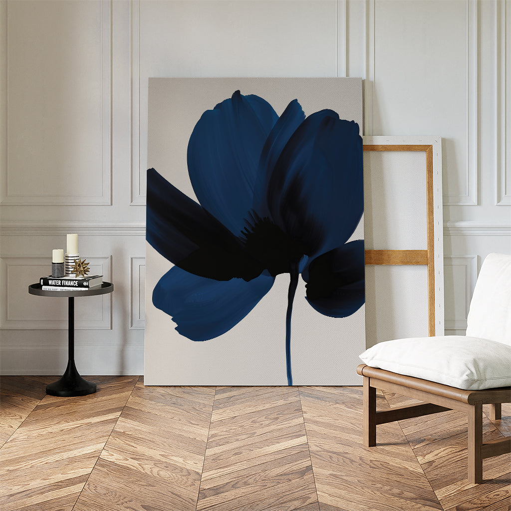The Nocturne Blossom Canvas Art, a large minimalist piece with a dark blue abstract flower, leans against a white paneled wall. An empty wooden frame, modern chair, and round table with candles and books accent the living room decor.