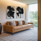 A modern living room with a tan leather sofa, four patterned cushions, and striking Noir Bloom Set of 2 wall art above features a patterned rug, tall vase, and a large window overlooking greenery.