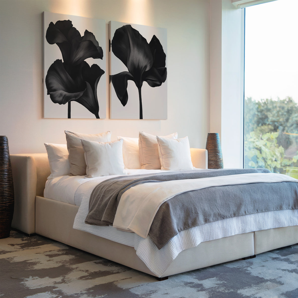A modern bedroom with a large bed featuring white and gray bedding, several pillows, Noir Bloom Set of 2 wall art above the headboard, and large windows letting in natural light.