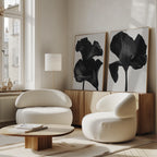 A modern living room with two white rounded armchairs, a wooden coffee table, a floor lamp, and the Noir Bloom Set of 2 wall art on a wooden sideboard against a light wall.
