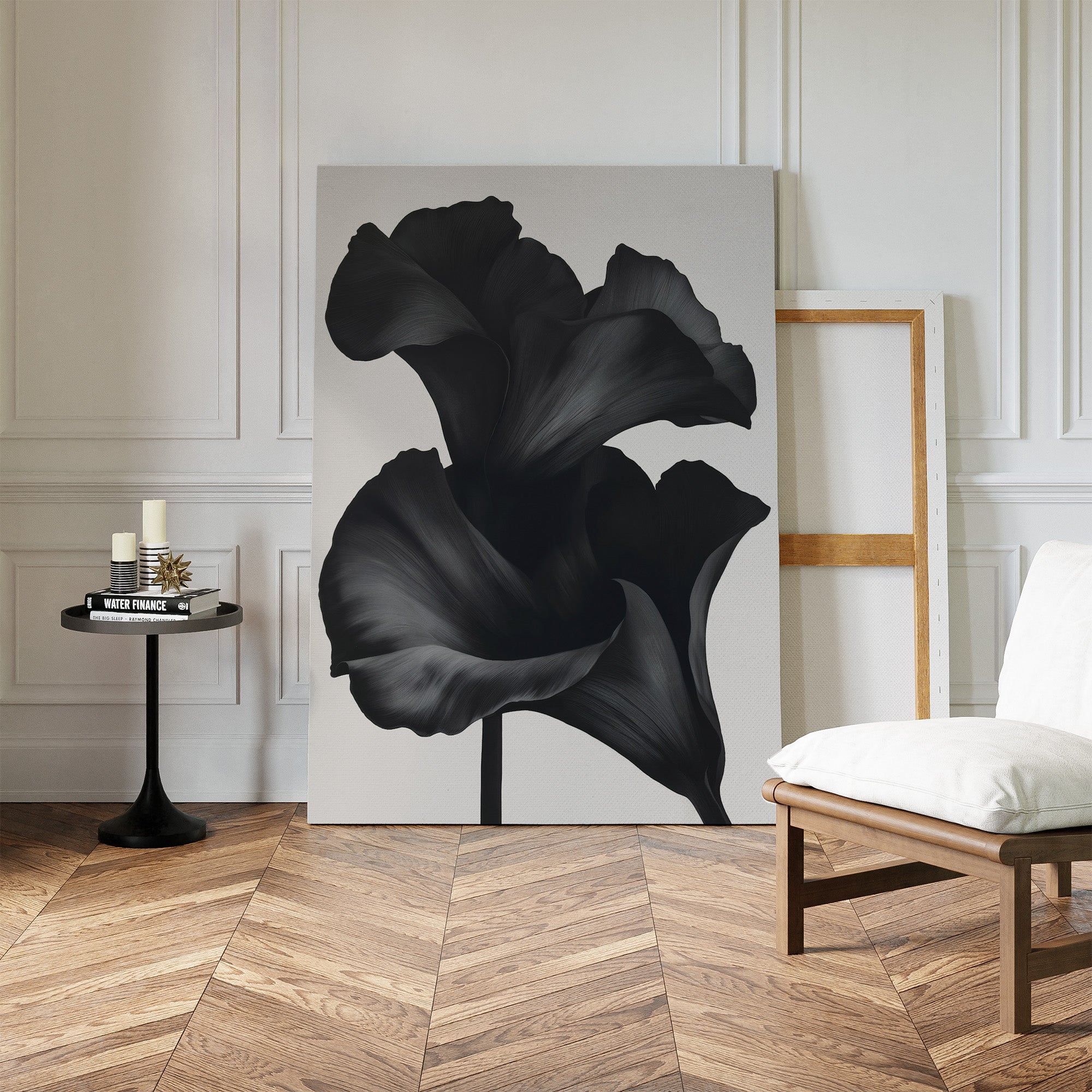 The Noir Bloom Abstract Canvas Art, a large minimalist piece with a black flower, leans against a white wall in an elegant room with herringbone floors, a cushioned chair, and a small round table displaying books and candles.
