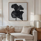 A modern living room features a beige sofa with neutral cushions, a side table with a lamp, and the Noir Bloom Canvas Art - Square above the sofa. A vase filled with dried flowers sits on the coffee table.