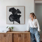 A woman in a white blouse and jeans leans against a wooden sideboard, admiring the Noir Bloom Canvas Art - Square, a striking minimalist piece, displayed in a modern living room with a potted plant and books.