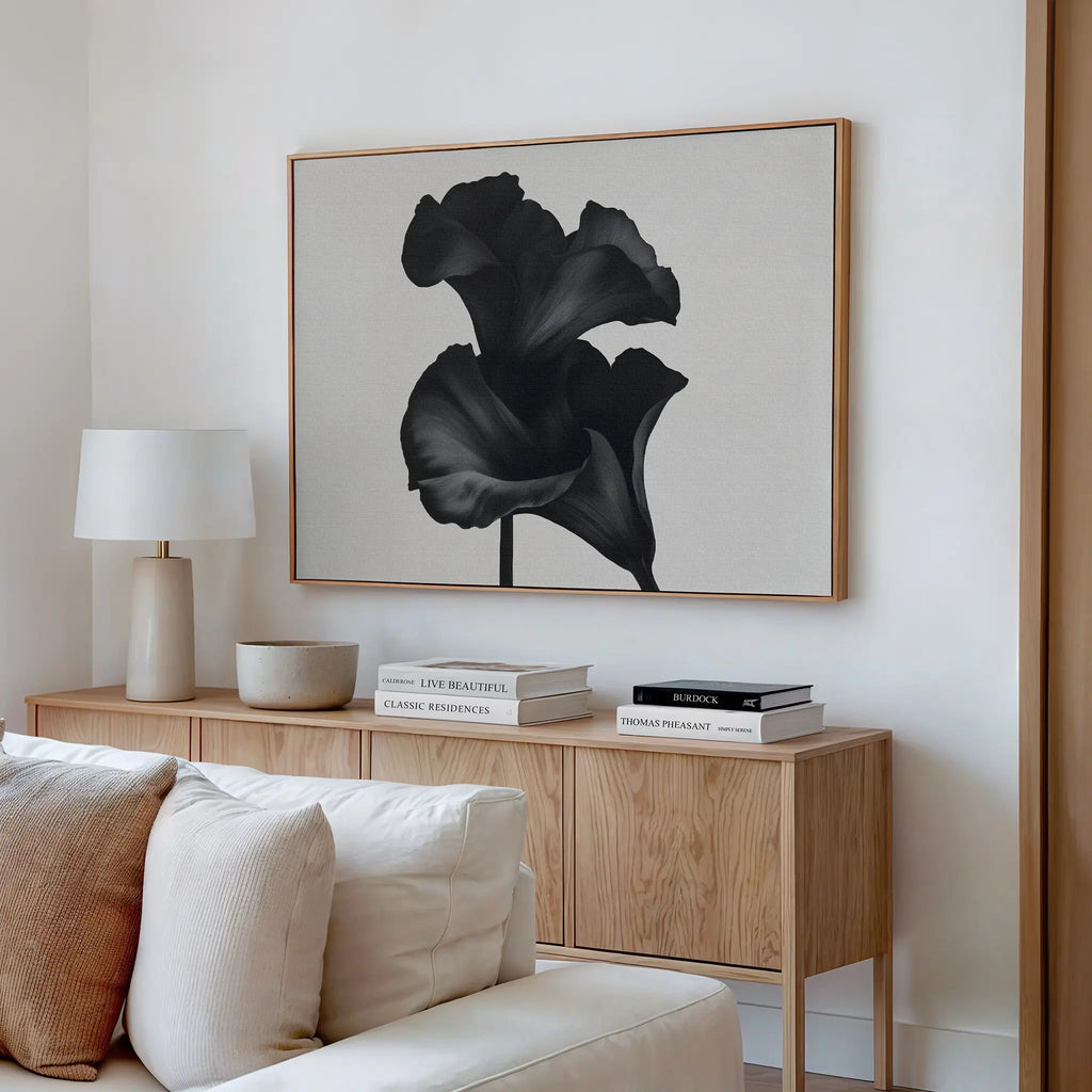 A modern living room with a light wood sideboard, neutral decor, white table lamp, and books. Hanging above is the Noir Bloom Canvas Art – Horizontal, featuring a black flower on a light background. A beige couch sits in the foreground.