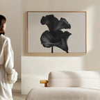 In a minimalistic room, a person in white admires the Noir Bloom Canvas Art – Horizontal, a large black flower artwork framed above a modern light sofa, highlighting refined living room decor.