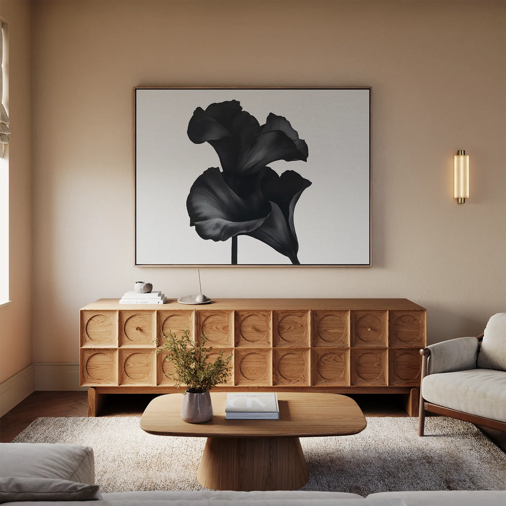 A modern living room with a light sofa, wooden coffee table, textured sideboard, and minimalist decor features the Noir Bloom Canvas Art – Horizontal above the sideboard, enhancing the space with its bold black-and-white floral design.