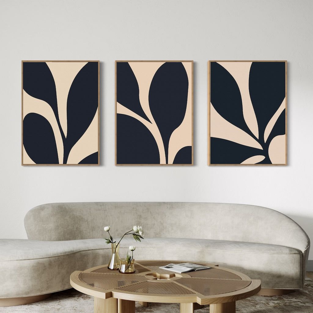 The Noir Blossom Set of 3 large wall art prints with beige and black organic shapes hangs above a curved, light sofa. A round wooden coffee table in front holds a vase with flowers and a book, creating a modern, minimal living room.