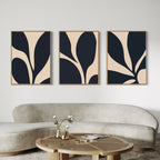 The Noir Blossom Set of 3 large wall art prints with beige and black organic shapes hangs above a curved, light sofa. A round wooden coffee table in front holds a vase with flowers and a book, creating a modern, minimal living room.