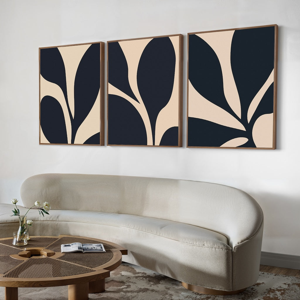 A modern living room with a curved beige sofa, round wooden coffee table, and the Noir Blossom Set of 3 minimalist wall art above the sofa. A small flower vase and a magazine on the table add a sleek, inviting touch.