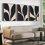 A modern living room features a beige sectional sofa, a round black coffee table, and the Noir Blossom Set of 3 minimalist botanical prints with black and beige shapes above the couch. A window shows greenery outside.