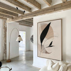 A bright, modern living space with wooden ceiling beams, a beige arc floor lamp, and white couch. The Noir Petals in Motion Abstract Canvas Art with black and beige shapes accents the white wall in this stunning design.
