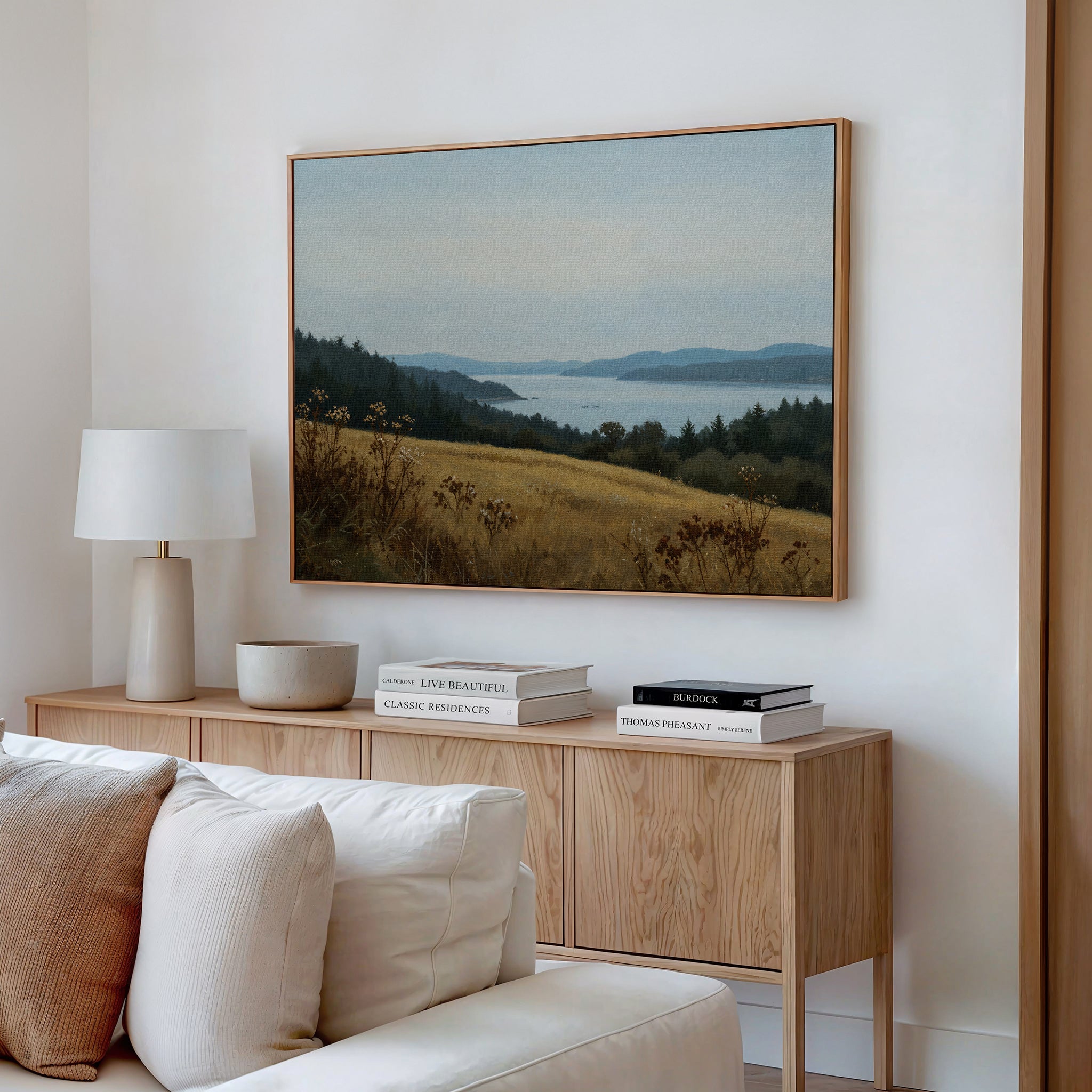A cozy, modern living room features a wooden sideboard with books, a bowl, and a white lamp. Above, the Northern Silence Canvas Art—a minimalist landscape of hills and water—enhances the decor. A white sofa with cushions sits in front.