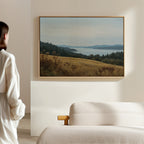 A person in a white shirt stands in a softly lit living room, gazing at the Northern Silence Canvas Art above a beige sofa; this minimalist wall art showcases rolling hills, trees, and a calm sky over distant water.