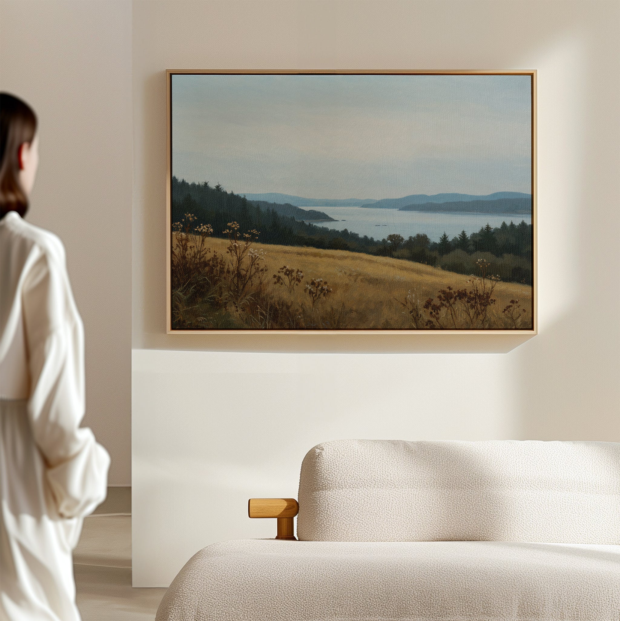 A person in a white shirt stands in a softly lit living room, gazing at the Northern Silence Canvas Art above a beige sofa; this minimalist wall art showcases rolling hills, trees, and a calm sky over distant water.