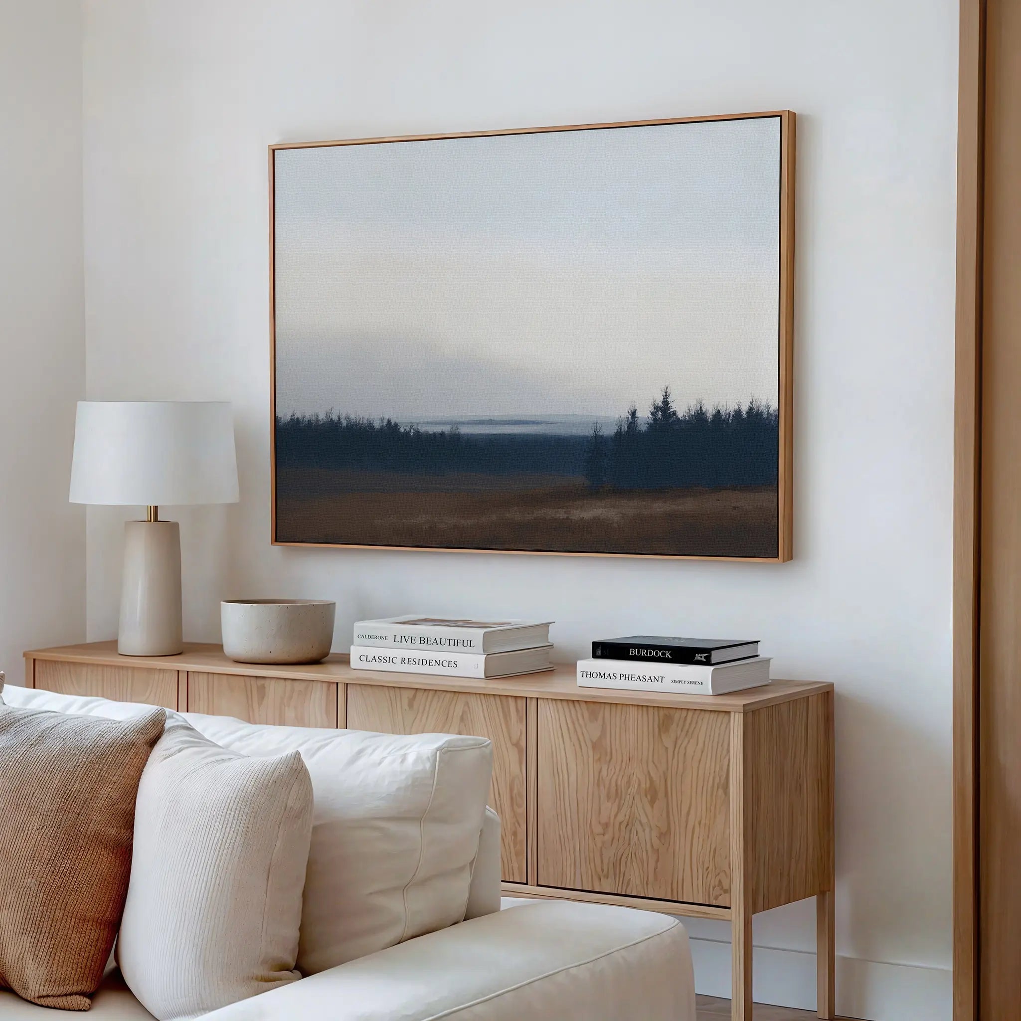 A minimalist living room with a light wood sideboard, white lamp, stacked books, a bowl, and the Northern Woods Canvas Art above a cream sofa evokes the tranquil beauty of a Nordic Park.