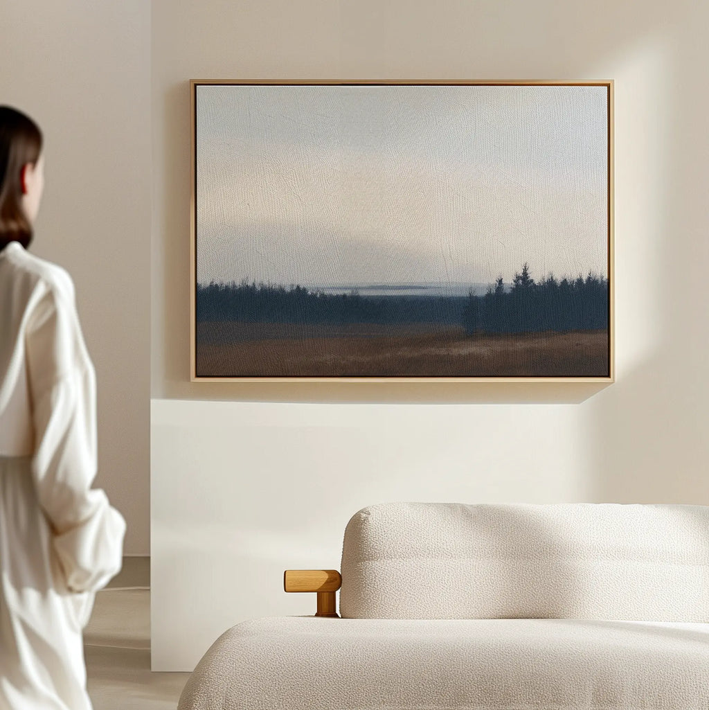 A person in a white robe stands in a minimalist room, gazing at the Northern Woods Canvas Art above a light-colored sofa. The artwork features a serene misty forest with trees and an overcast sky.