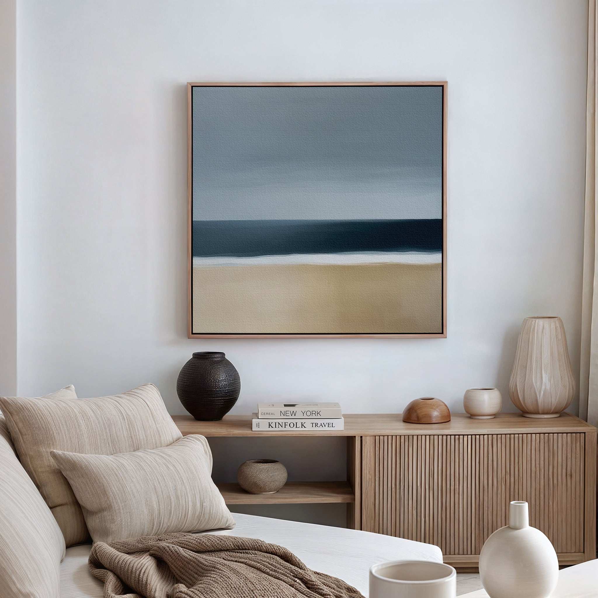 A minimal living room with a beige sofa, striped cushions, and modern wooden sideboard, topped with books and vases. Above hangs the Ocean Edge Canvas Art in muted blue, beige, and cream tones for a calm coastal vibe.