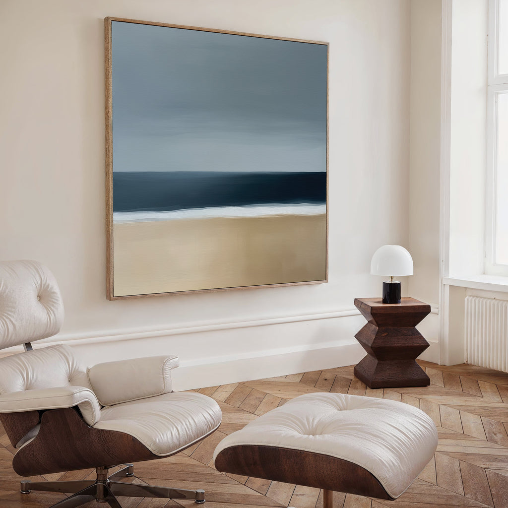 A modern, minimalist room with a white leather lounge chair, ottoman, small wooden side table with a dome-shaped lamp, herringbone flooring, and the striking Ocean Edge Canvas Art adds sophistication to the serene space.