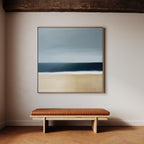A minimalist interior showcases the Ocean Edge Canvas Art—a coastal beach scene—hung above a simple wooden bench with a brown leather cushion, all set against a light-colored wall and wooden floor.