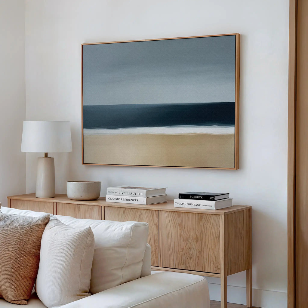 A minimalist living room features a wooden sideboard with stacked books, a lamp, and a ceramic bowl, while the Ocean Edge Canvas Art – Horizontal hangs above a cream-colored sofa on a white wall.