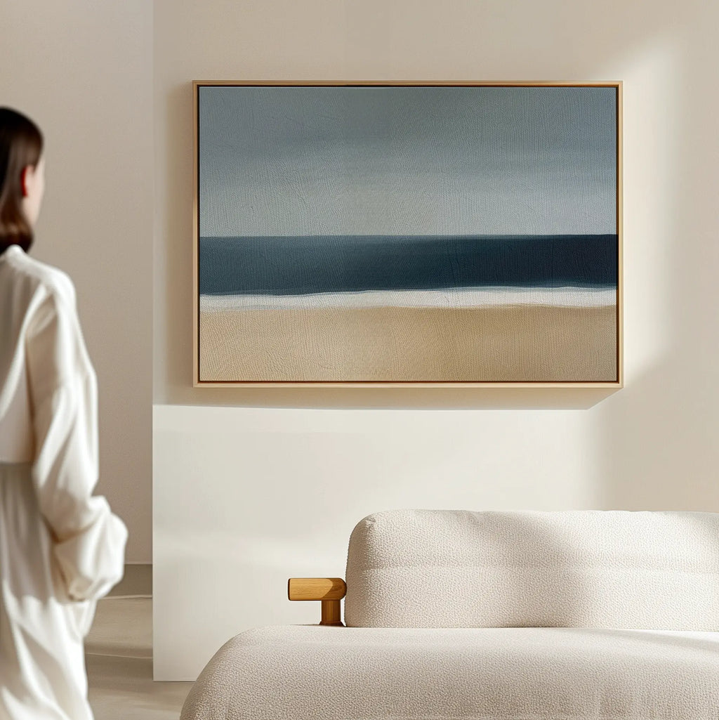 A person in a white robe stands in a minimal, light-filled room with a beige sofa and the Ocean Edge Canvas Art – Horizontal, featuring an abstract beach scene, framed on the wall.