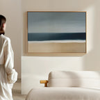 A person in a white robe stands in a minimal, light-filled room with a beige sofa and the Ocean Edge Canvas Art – Horizontal, featuring an abstract beach scene, framed on the wall.