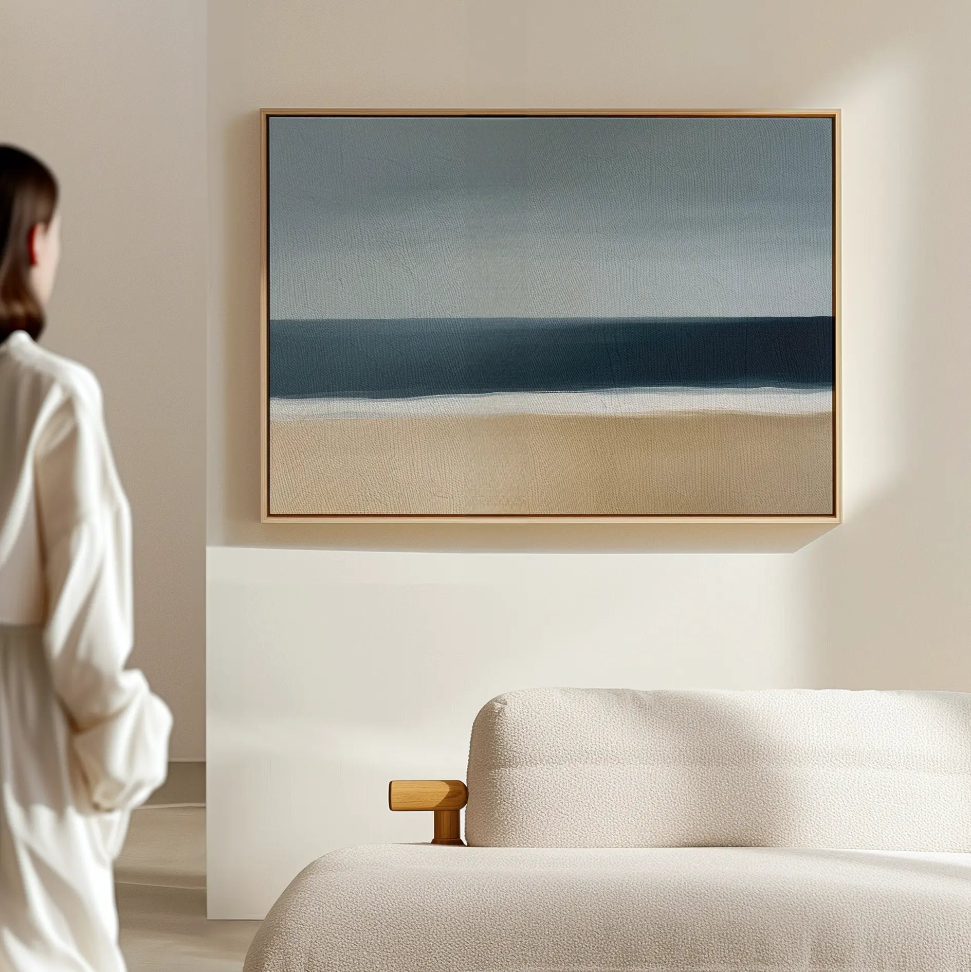 A person in a white robe stands in a minimal, light-filled room with a beige sofa and the Ocean Edge Canvas Art – Horizontal, featuring an abstract beach scene, framed on the wall.