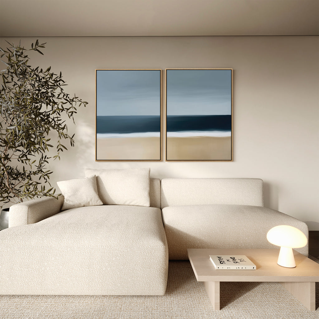 A modern living room with a cream sectional sofa, wooden coffee table, indoor plant, and the Ocean Edge Set of 2 coastal wall art above the couch adds a touch of elegant decor.