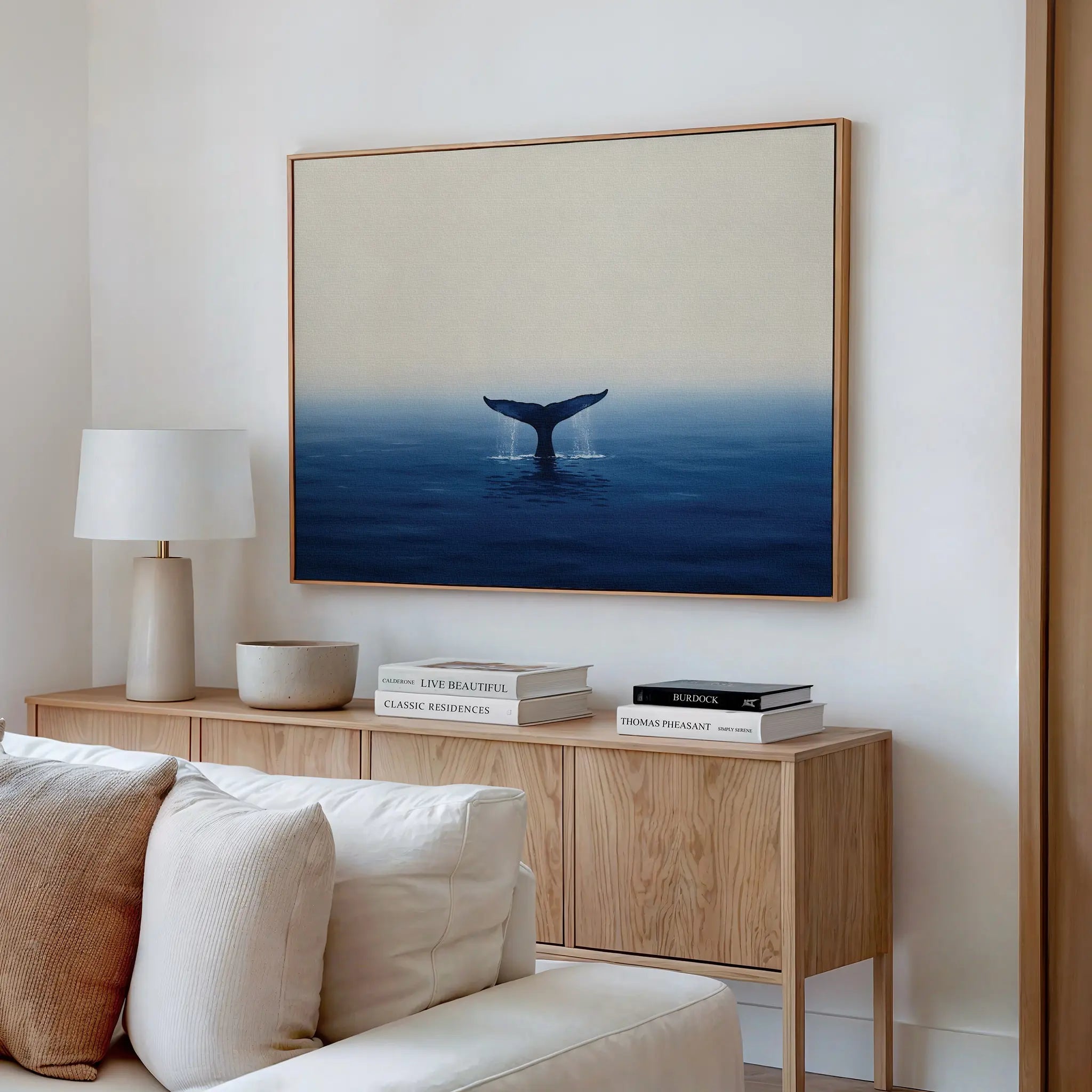 A modern living room showcases the Ocean Giant Canvas Art of a whale above blue water on a white wall, joined by a light wood sideboard, white lamp, books, and a beige sofa.