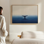 A woman in a white outfit stands in a minimalist room, admiring the Ocean Giant Canvas Art—whale tail rising from a tranquil blue sea—displayed on a light wall above a modern sofa.