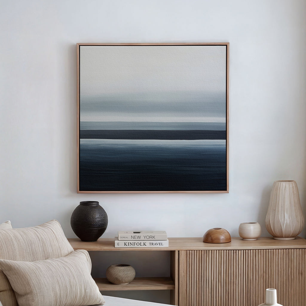 A minimalist living space with neutral decor, featuring a wooden console table with ceramics and books, and the Oceanic Blur Abstract Canvas Art with blue and gray gradients displayed on a white wall above.