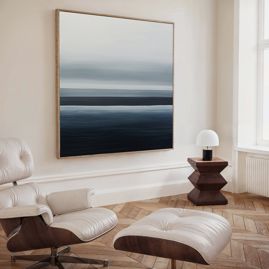 A modern minimalist living room features a white leather lounge chair with ottoman, a geometric wood side table with a white lamp, and the Oceanic Blur Abstract Canvas Art above a light wood herringbone floor.