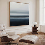 A modern minimalist living room features a white leather lounge chair with ottoman, a geometric wood side table with a white lamp, and the Oceanic Blur Abstract Canvas Art above a light wood herringbone floor.