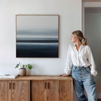 A woman in a white blouse and jeans stands by a wooden sideboard, admiring the Oceanic Blur Abstract Canvas Art—a minimalist piece in blue and gray tones, ideal for coastal living room decor. A small plant and book rest on the sideboard.