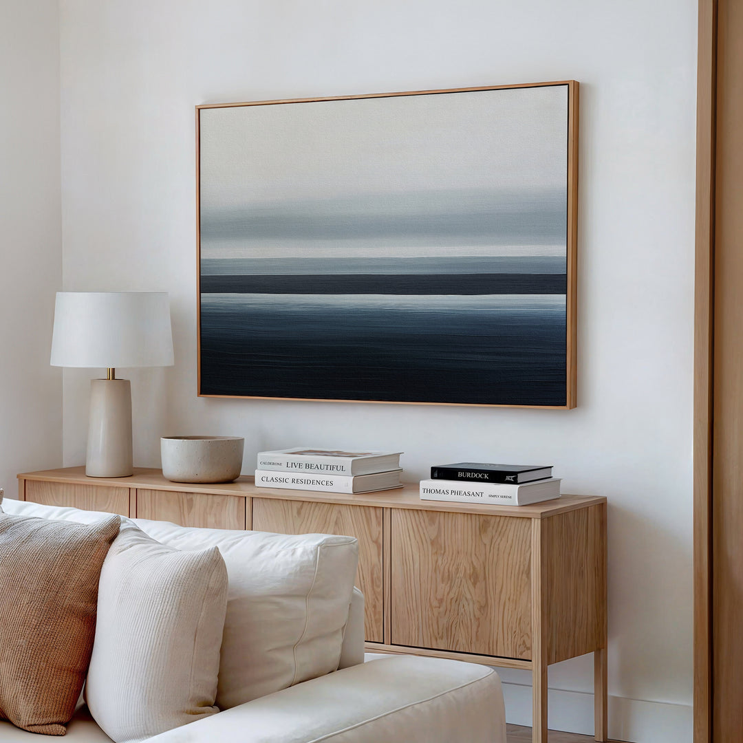 A modern living room features the Oceanic Blur Canvas Art – Horizontal in cool tones above a light sofa, paired with a light wood sideboard, minimalist decor, white lamp, stacked books, and a bowl.