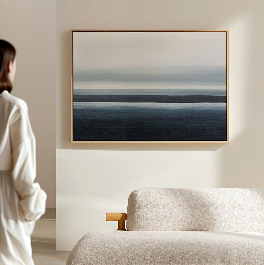A woman in a light outfit stands in a modern room with a beige sofa and the Oceanic Blur Canvas Art – Horizontal, an abstract painting of blue and gray stripes that adds to the minimalist decor and subtle coastal vibe.