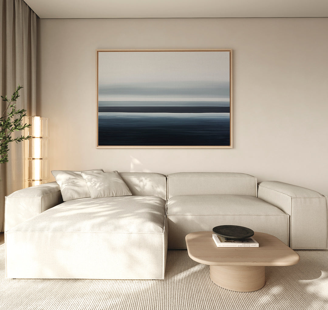 A modern living room featuring a cream sectional, light wood coffee table with a dark bowl, beige rug, tall lamp, potted plant, and the Oceanic Blur Canvas Art – Horizontal creating a coastal vibe with its abstract blue tones.