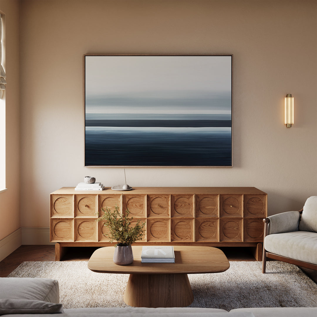 A minimalist living room features a wooden sideboard, the Oceanic Blur Canvas Art – Horizontal above it, a small coffee table with books and a vase, a cozy rug, and a light sofa. Warm neutral tones create an inviting atmosphere.