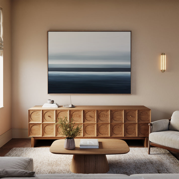 A minimalist living room features a wooden sideboard, the Oceanic Blur Canvas Art – Horizontal above it, a small coffee table with books and a vase, a cozy rug, and a light sofa. Warm neutral tones create an inviting atmosphere.