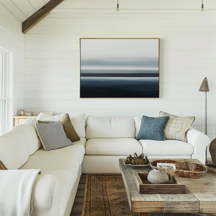A cozy living room with minimalist decor features a white sectional, neutral and blue pillows, a rustic wooden coffee table, and the Oceanic Blur Canvas Art – Horizontal for a coastal vibe. Natural light enhances the white shiplap walls.