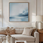 A modern living room with a beige sofa, neutral pillows, a light wood side table with a white lamp, dried flowers in a vase, and the Oceanic Overcast Canvas Art above the sofa creates a calming atmosphere.