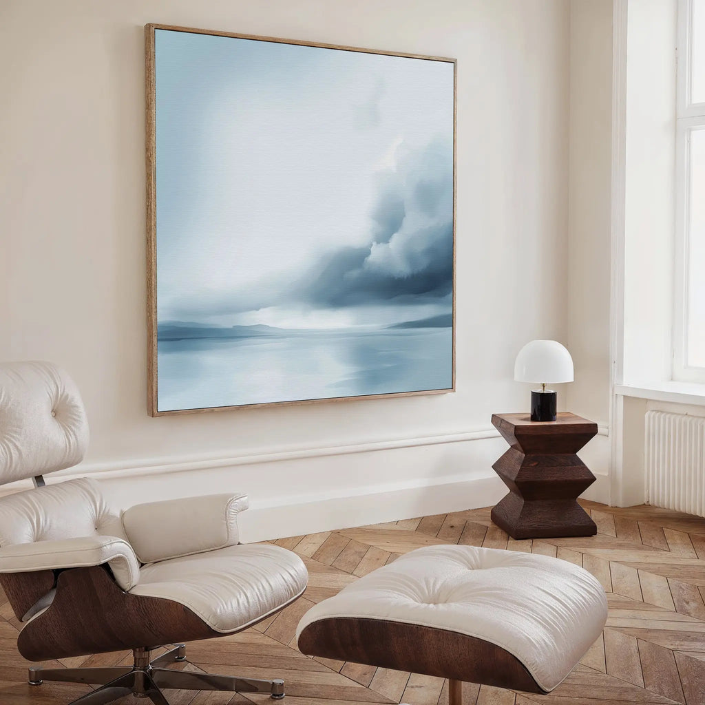 A modern, minimalist living room features the Oceanic Overcast Canvas Art as wall décor, a white leather lounge chair with ottoman, a geometric dark wood side table, and a small white lamp by a window with natural light.