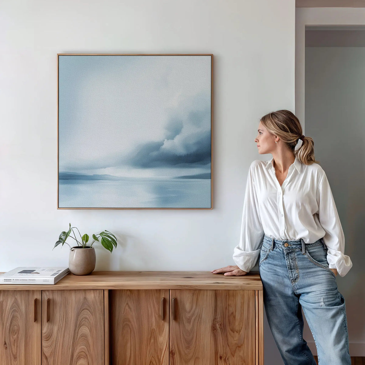 A woman in a white blouse and jeans leans against a wooden cabinet, gazing at the Oceanic Overcast Canvas Art hanging on the white wall above her. A potted plant and books add to the tranquil mood of the space.