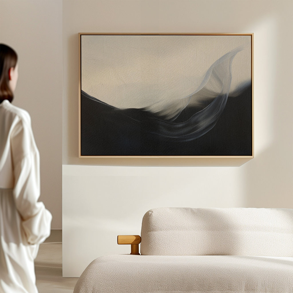 A person stands in a minimal, modern room with a light sofa and wooden armrest, admiring the Oceanic Pulse Abstract Canvas Art—an abstract painting with dark and light flowing shapes—hanging as stylish living room decor on a beige wall.