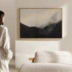 A person stands in a minimal, modern room with a light sofa and wooden armrest, admiring the Oceanic Pulse Abstract Canvas Art—an abstract painting with dark and light flowing shapes—hanging as stylish living room decor on a beige wall.