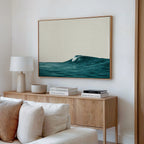 A modern living room features a wooden sideboard with books, a lamp, and a ceramic bowl. Above it hangs the Oceanside Crest Canvas Art, its ocean wave design complementing the white sofa and coastal decor.